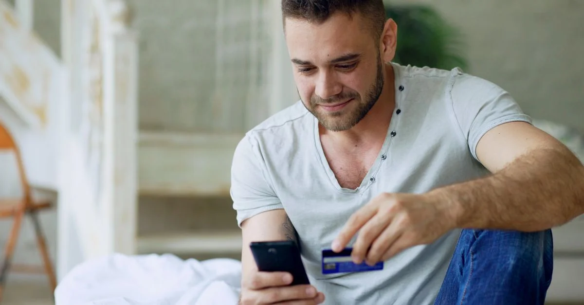 Homme souriant utilisant un smartphone pour faire des achats en ligne dans un cadre intérieur confortable.